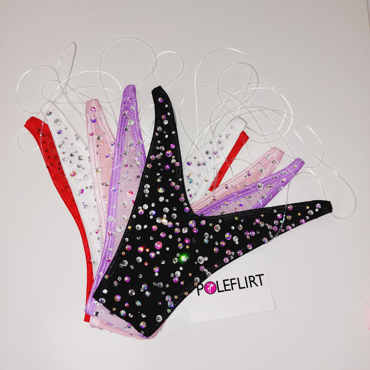 Five pole dance pole thong bikini bottoms in red, white, pink, lavender and black, embellished with rhinestones and zippers, arranged on a white background with a 'POLEFLIRT' brand tag.