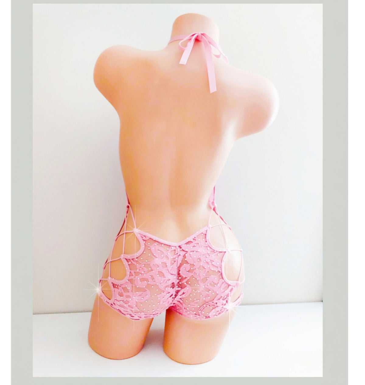 Back view of a Back view of a beige mannequin torso displaying a baby pink lace one piece short set with rhinestone embellishments and side lcord lace ups.
