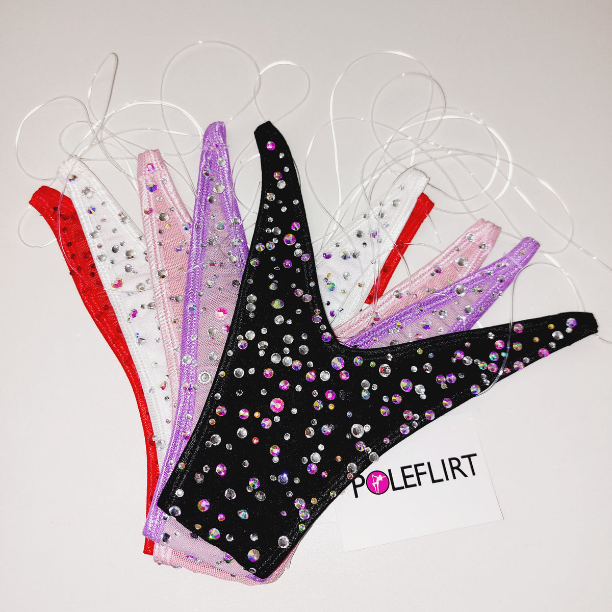 Four women's pole dance thongs with clear elastic straps against a light gray background. The bikini bottoms are shown in red, white, pink, and black, all embellished with iridescent rhinestones.