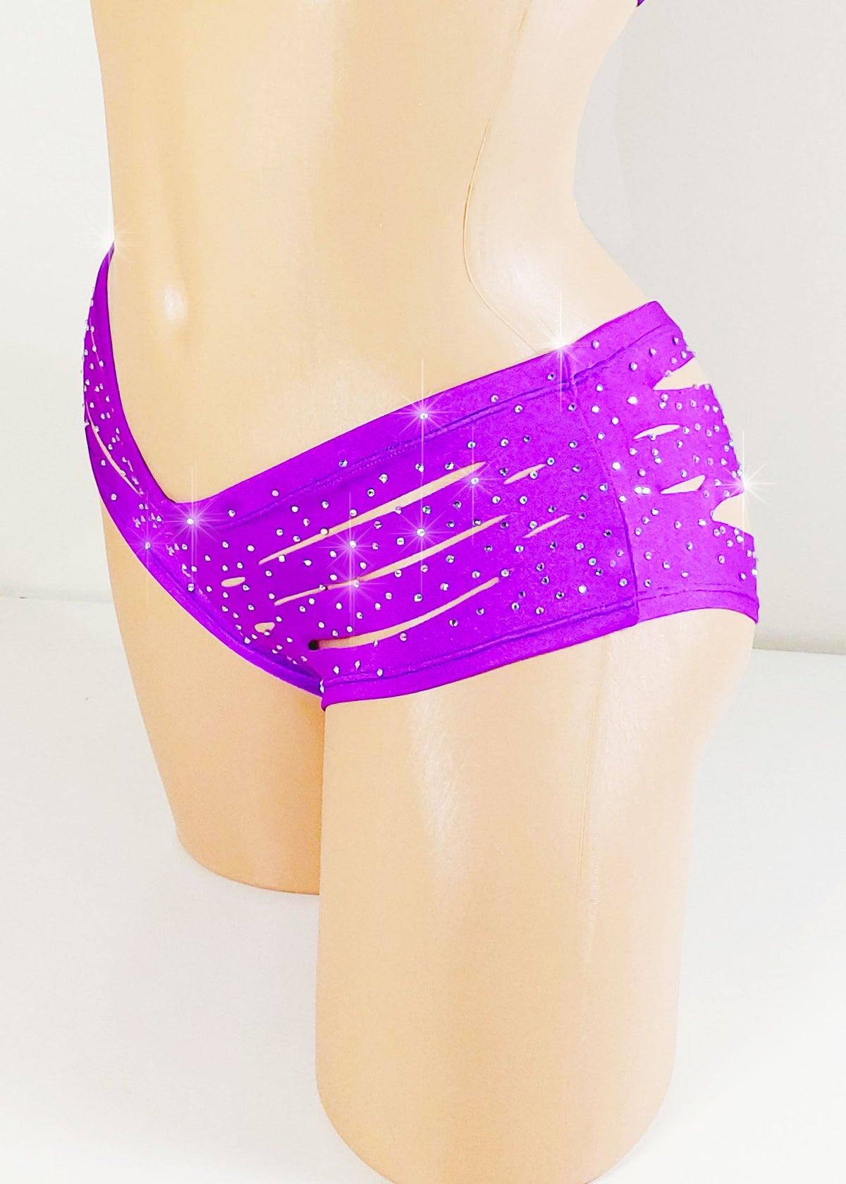 Purple shorts with multiple horizontal cutouts and accented with sparklling rhinestones on a plastic beige mannequin torso against a plain background