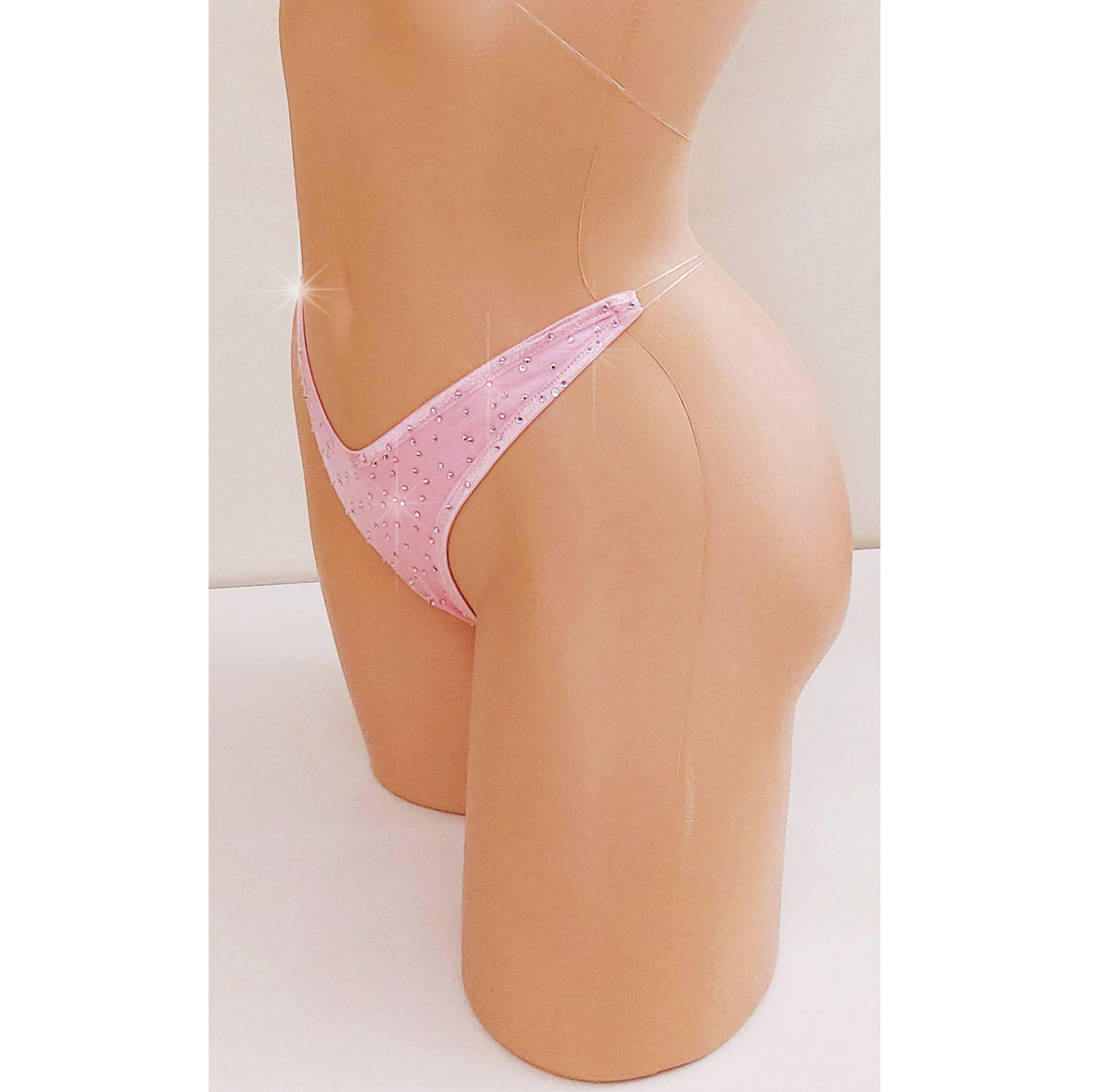 A close-up side view of a mannequin torso wearing a light pink thong panties that are covered in small rhinestones and have clear straps that appear invisible and transparent, displayed on a beige mannequin torso against a neutral light background.