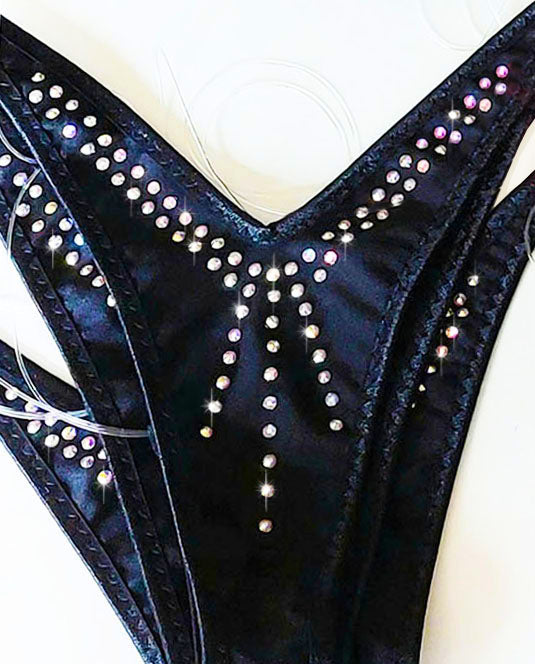 Close-up of a bunch of black bikini bottoms decorated with rows of iridescent rhinestones and crystals, set against a plain light background.
