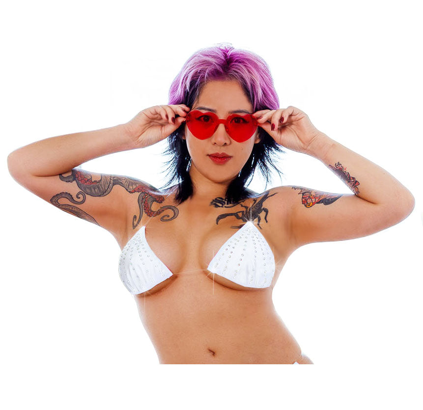 A woman with short purple hair and arm tattoos wears a white triangle bikini top and red heart-shaped sunglasses against a plain white background.